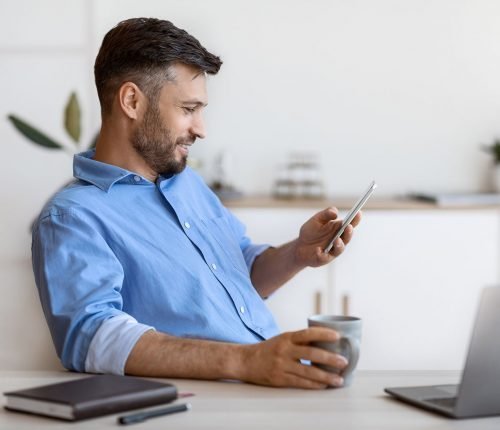 Handsome Male Freelancer Using Smartphone And Having Coffee At Home Office, Enjoying Break At Work, Businessman Browsing Internet Or Messaging With Friends, Sitting At Desk With Laptop, Copy Space